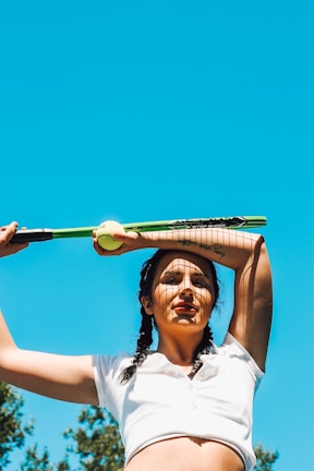 A person with braided hair is holding a tennis racket and a ball above their head. They are wearing a white shirt and standing under a clear blue sky with some trees in the background.