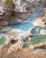 A serene natural hot spring setting with three individuals relaxing in different small pools of warm, mineral-rich water. The scene is surrounded by rocks, and in the background, a small stream flows through a snowy, forested area.