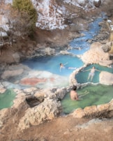 A serene natural hot spring setting with three individuals relaxing in different small pools of warm, mineral-rich water. The scene is surrounded by rocks, and in the background, a small stream flows through a snowy, forested area.