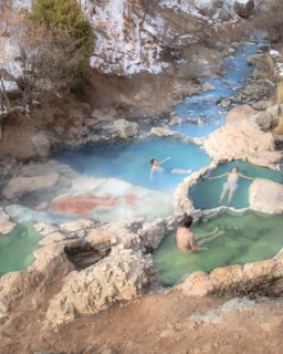 A serene natural hot spring setting with three individuals relaxing in different small pools of warm, mineral-rich water. The scene is surrounded by rocks, and in the background, a small stream flows through a snowy, forested area.