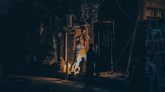 A person wearing striped clothing is working on a wooden surface mounted vertically, possibly hammering or adjusting it. The scene is illuminated by a warm light source, creating dramatic shadows. Two statues and some construction materials are visible in the background. The setting is dark, possibly an outdoor space or an unfinished room.