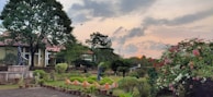 A tranquil garden landscape featuring lush greenery, a variety of flowering plants, and a few sculptures strategically placed among the trees and bushes. A path lined with potted plants adds structure to the scene, while the sky displays a subdued sunset with clouds.
