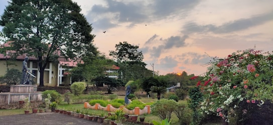 A tranquil garden landscape featuring lush greenery, a variety of flowering plants, and a few sculptures strategically placed among the trees and bushes. A path lined with potted plants adds structure to the scene, while the sky displays a subdued sunset with clouds.