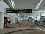 A spacious airport terminal corridor featuring a high, curved ceiling with large windows for natural light. Signs for international departures, immigration, and health services are prominently displayed. People are seen walking and pulling luggage, and a coffee shop is located to the left. The area has a modern design with patterned flooring.