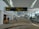 A spacious airport terminal corridor featuring a high, curved ceiling with large windows for natural light. Signs for international departures, immigration, and health services are prominently displayed. People are seen walking and pulling luggage, and a coffee shop is located to the left. The area has a modern design with patterned flooring.