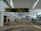 A spacious airport terminal corridor featuring a high, curved ceiling with large windows for natural light. Signs for international departures, immigration, and health services are prominently displayed. People are seen walking and pulling luggage, and a coffee shop is located to the left. The area has a modern design with patterned flooring.