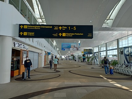 A spacious airport terminal corridor featuring a high, curved ceiling with large windows for natural light. Signs for international departures, immigration, and health services are prominently displayed. People are seen walking and pulling luggage, and a coffee shop is located to the left. The area has a modern design with patterned flooring.