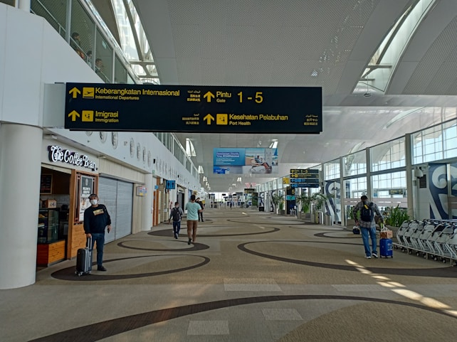 A spacious airport terminal corridor featuring a high, curved ceiling with large windows for natural light. Signs for international departures, immigration, and health services are prominently displayed. People are seen walking and pulling luggage, and a coffee shop is located to the left. The area has a modern design with patterned flooring.