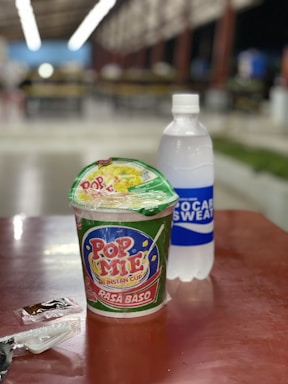 A cup of instant noodles labeled 'Pop Mie' sits on a table next to a bottle of Pocari Sweat. Several small packets, possibly seasoning or utensils, are also present on the table. The background appears to be a blurred indoor setting with tables and lights.