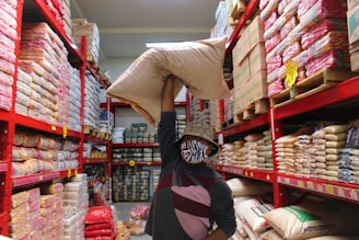 A person is holding a large sack over their shoulder while standing in a grocery or storage aisle lined with shelves full of various packaged goods. The individual is wearing a patterned bucket hat and a face mask, and the shelves are organized with many similar items stacked closely together. Bright red metal frames support the stocked shelves and signage is visible indicating prices or product information.