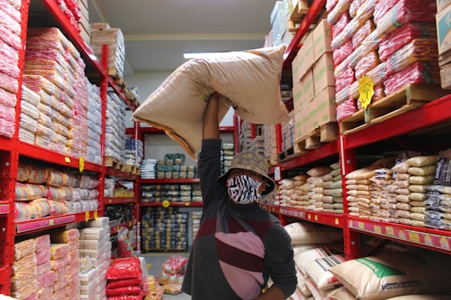 A person is holding a large sack over their shoulder while standing in a grocery or storage aisle lined with shelves full of various packaged goods. The individual is wearing a patterned bucket hat and a face mask, and the shelves are organized with many similar items stacked closely together. Bright red metal frames support the stocked shelves and signage is visible indicating prices or product information.