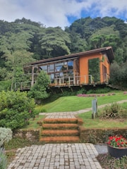 A modern wooden house with large glass windows is nestled among lush greenery and trees. The house features an orange wall and is elevated on a well-manicured grassy hill with stairs leading up to it. Decorative plants and flowers surround the area, enhancing the natural setting.