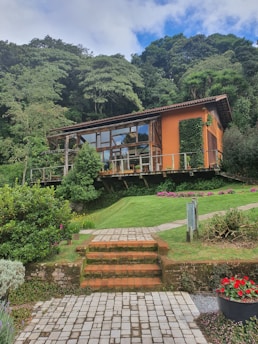 A modern wooden house with large glass windows is nestled among lush greenery and trees. The house features an orange wall and is elevated on a well-manicured grassy hill with stairs leading up to it. Decorative plants and flowers surround the area, enhancing the natural setting.