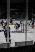 A group of ice hockey players in dark blue uniforms is on the ice at a rink. The scene shows a goalie with protective gear near the goal post, while other players are positioned around him. The background includes empty spectator seats and glass panels around the rink.