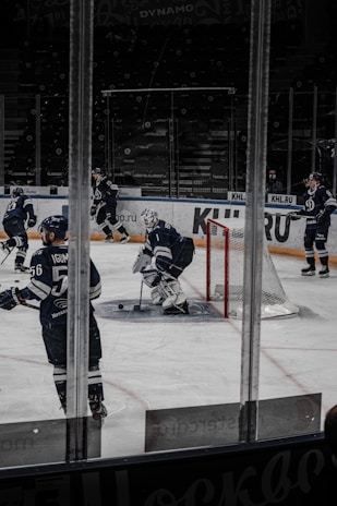 A group of ice hockey players in dark blue uniforms is on the ice at a rink. The scene shows a goalie with protective gear near the goal post, while other players are positioned around him. The background includes empty spectator seats and glass panels around the rink.