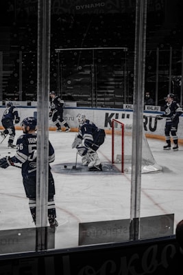 A group of ice hockey players in dark blue uniforms is on the ice at a rink. The scene shows a goalie with protective gear near the goal post, while other players are positioned around him. The background includes empty spectator seats and glass panels around the rink.