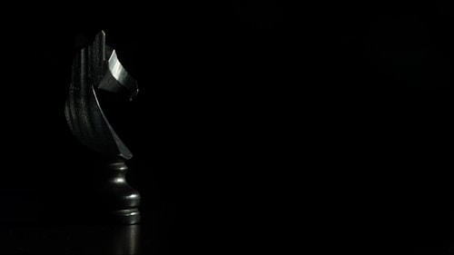 A black chess knight piece is dramatically lit against a dark background, highlighting its smooth and polished surface. The piece is positioned to the left side of the frame, creating a sense of mystery and focus on the intricately carved details.