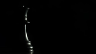 A chess queen is placed prominently on the left side, emerging from the shadows with a metallic sheen against a dark, minimalist background.