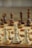 A close-up view of a chessboard with wooden chess pieces arranged in starting positions, featuring pawns, knights, bishops, rooks, a queen, and a king in focus. The foreground pieces are made of light wood while the background pieces are darker, out of focus.