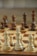 A close-up view of a chessboard with wooden chess pieces arranged in starting positions, featuring pawns, knights, bishops, rooks, a queen, and a king in focus. The foreground pieces are made of light wood while the background pieces are darker, out of focus.