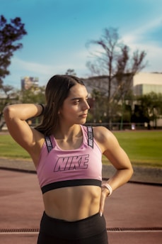 A person standing outdoors on a sunny day, wearing a pink Nike sports bra and black leggings. The individual has their hand resting on the back of their head, looking confidently into the distance. In the background, there are trees, a building, and a blue sky.