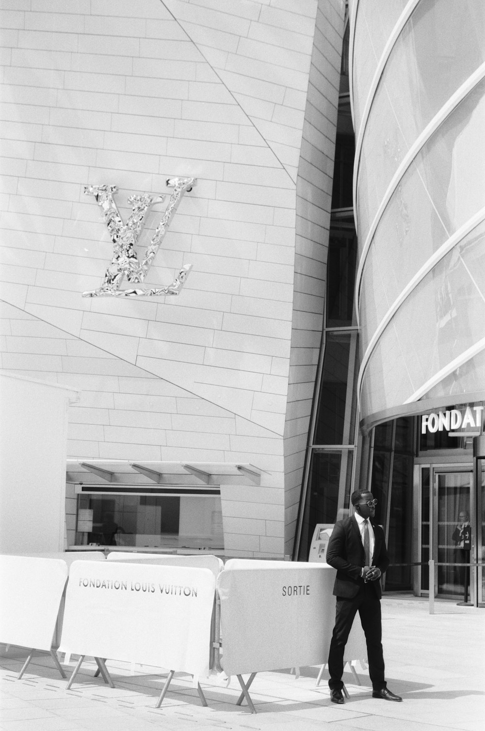 grayscale photo of man in black jacket standing beside building