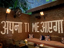 A cozy outdoor seating area with a rustic brick wall featuring large white text in a mixture of Hindi and English. The area is warmly lit by wall-mounted lamps, and there are leafy green plants visible above the wall. The seating consists of a cushion-covered bench with patterned pillows and a table with dining chairs.
