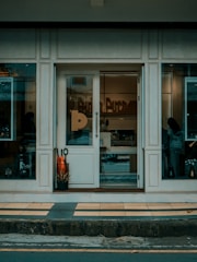 The storefront has a glass door surrounded by large windows with a neutral color scheme. Inside, there are decorative items and a silhouette of a person is visible. There are two large orange plants in a black pot near the entrance.