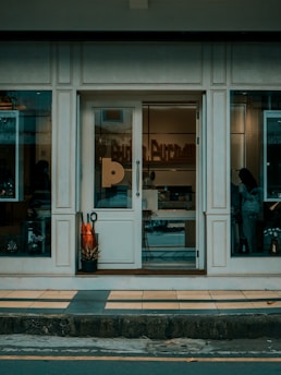 The storefront has a glass door surrounded by large windows with a neutral color scheme. Inside, there are decorative items and a silhouette of a person is visible. There are two large orange plants in a black pot near the entrance.
