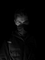 A person is partially visible in low light, wearing dark clothing and a mask that covers the nose and mouth, leaving only the eyes exposed. The background is completely black, enhancing the mysterious and shadowy appearance.