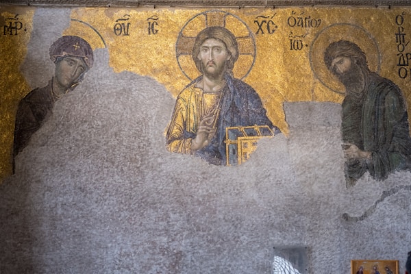 A Byzantine mosaic depicting religious figures adorned with halos. The figures on either side have inscriptions near them, featuring intricate and ornate detailing. The central figure holds a book and is set against a gilded backdrop. The mosaic is partially eroded, revealing worn textures in some areas.