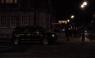 A dark street scene at night featuring a black taxi parked by a building with illuminated windows. The Battersea Arts Centre is visible in the background, with its sign glowing in yellow. Street lamps cast a warm light over the sidewalk where a few people are walking.