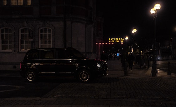 A dark street scene at night featuring a black taxi parked by a building with illuminated windows. The Battersea Arts Centre is visible in the background, with its sign glowing in yellow. Street lamps cast a warm light over the sidewalk where a few people are walking.