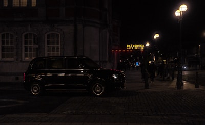 A dark street scene at night featuring a black taxi parked by a building with illuminated windows. The Battersea Arts Centre is visible in the background, with its sign glowing in yellow. Street lamps cast a warm light over the sidewalk where a few people are walking.