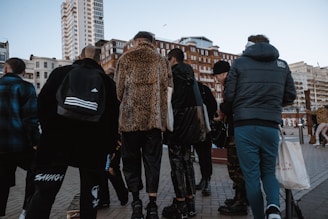 A group of people stands together on a city street. They are wearing various styles of clothing, including a leopard print coat and a sporty jacket. Skyscrapers and older architectural buildings loom in the background, suggesting an urban setting.