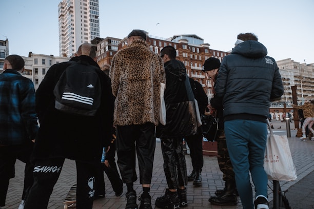 A group of people stands together on a city street. They are wearing various styles of clothing, including a leopard print coat and a sporty jacket. Skyscrapers and older architectural buildings loom in the background, suggesting an urban setting.