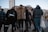 A group of people stands together on a city street. They are wearing various styles of clothing, including a leopard print coat and a sporty jacket. Skyscrapers and older architectural buildings loom in the background, suggesting an urban setting.
