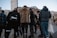A group of people stands together on a city street. They are wearing various styles of clothing, including a leopard print coat and a sporty jacket. Skyscrapers and older architectural buildings loom in the background, suggesting an urban setting.