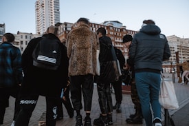 A group of people stands together on a city street. They are wearing various styles of clothing, including a leopard print coat and a sporty jacket. Skyscrapers and older architectural buildings loom in the background, suggesting an urban setting.