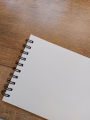 A spiral-bound notebook with a blank white page is placed on a wooden surface. The notebook is positioned diagonally, showcasing its metal binding rings on the left side.