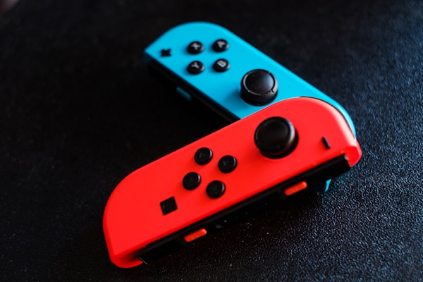 Two colorful game controllers, one red and one blue, are positioned on a dark textured surface. Each controller features several buttons and joysticks, emphasizing their functionality.