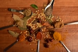 A variety of spices are artfully arranged on a wooden surface. There are several spoons containing different spices such as turmeric, cumin seeds, chili powder, and coriander seeds. Other spices like bay leaves, cardamom pods, and dried peppers are scattered across the table, creating a vibrant and textured display.