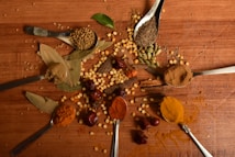 A variety of spices are artfully arranged on a wooden surface. There are several spoons containing different spices such as turmeric, cumin seeds, chili powder, and coriander seeds. Other spices like bay leaves, cardamom pods, and dried peppers are scattered across the table, creating a vibrant and textured display.