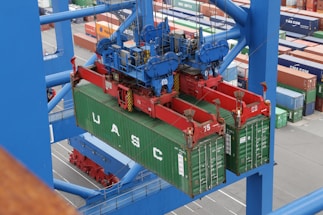 A large overhead crane is lifting a green shipping container in a busy port terminal. The crane is blue with red mechanical components, and the container is marked with the letters UASC. Numerous stacked shipping containers in various colors are visible in the background, showing organization and logistics.