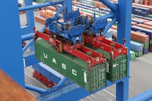 A large overhead crane is lifting a green shipping container in a busy port terminal. The crane is blue with red mechanical components, and the container is marked with the letters UASC. Numerous stacked shipping containers in various colors are visible in the background, showing organization and logistics.