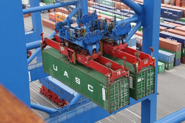 A large overhead crane is lifting a green shipping container in a busy port terminal. The crane is blue with red mechanical components, and the container is marked with the letters UASC. Numerous stacked shipping containers in various colors are visible in the background, showing organization and logistics.