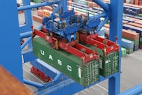 A large overhead crane is lifting a green shipping container in a busy port terminal. The crane is blue with red mechanical components, and the container is marked with the letters UASC. Numerous stacked shipping containers in various colors are visible in the background, showing organization and logistics.