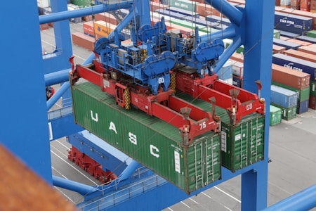 A large overhead crane is lifting a green shipping container in a busy port terminal. The crane is blue with red mechanical components, and the container is marked with the letters UASC. Numerous stacked shipping containers in various colors are visible in the background, showing organization and logistics.