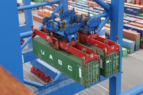 A large overhead crane is lifting a green shipping container in a busy port terminal. The crane is blue with red mechanical components, and the container is marked with the letters UASC. Numerous stacked shipping containers in various colors are visible in the background, showing organization and logistics.