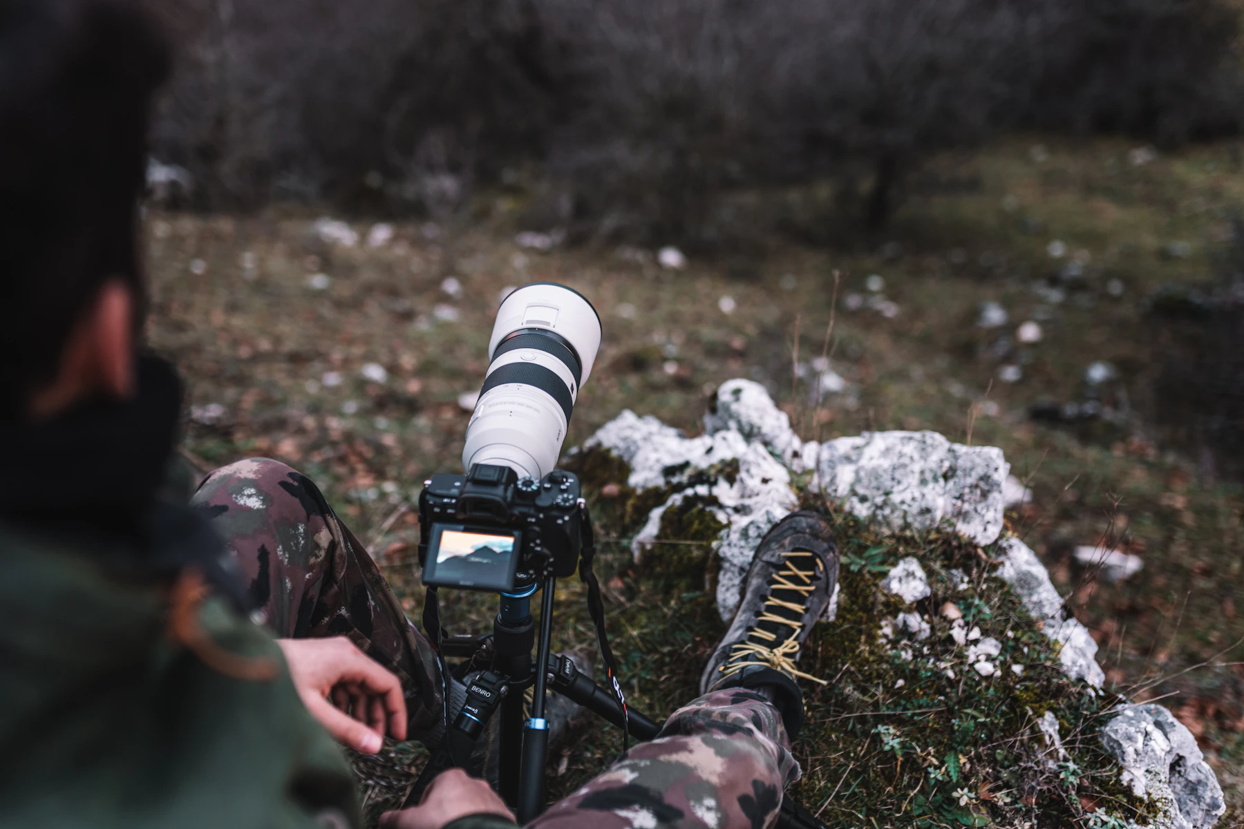 Seen over the shoulder: a white telephoto lens on a tripod in front of a seated photographer in camouflage trousers